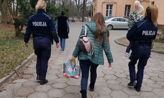 dwie umundurowane policjantki, w tym jedna niesie na ręku dziecko, między nimi idzie matka dziecka