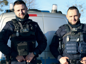 dwaj umundurowani policjanci stoją przed radiowozem