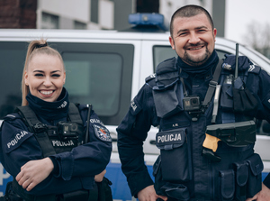 dwoje umundurowanych policjantów stoi przed radiowozem