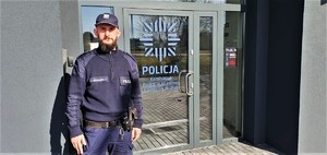 Zdjęcie kolorowe: umundurowany policjant, w tle widoczne drzwi do komisariatu