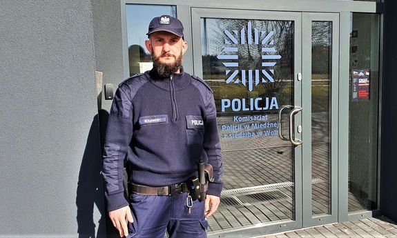 Zdjęcie kolorowe: umundurowany policjant, w tle widoczne drzwi do komisariatu