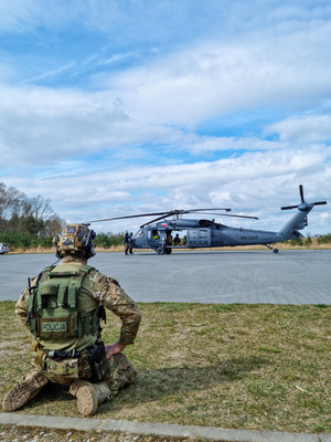 Na trawie, przed płytą lądowiska klęczy, funkcjonariusz Policji w mundurze taktycznym na jego głowie hełm, w tle śmigłowiec Black Hawk podczas rozruchu silników.