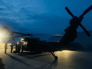 Nocne zdjęcie lądowiska śmigłowców, na płycie policyjny Black Hawk, przed lewymi drzwiami maszyny kilka osób w słabym oświetleniu pobliskiej latarni.