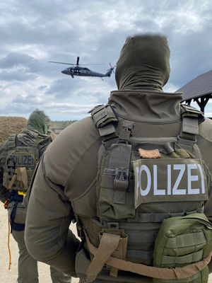 Dwaj funkcjonariusze niemieckiej Policji w mundurach taktycznych, sfotografowani od tyłu, przyglądają się przelatującemu Black Hawkowi.