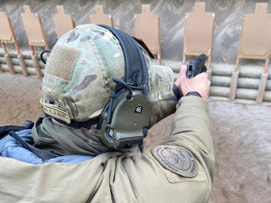 Strzelnica, z oddali i z zza pleców policjanta. W wyprostowanych rękach niemieckiego policjanta pistolet skierowany w stronę tarczy strzeleckiej, na ramieniu naszywka z napisem Stadtpolizei Zurich i grafika skorpiona.