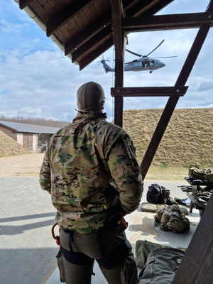 Pod wiatą, na tle wału ziemnego, funkcjonariusz w umundurowaniu taktycznym przyglądający się przelatującemu śmigłowcowi Black Hawk.