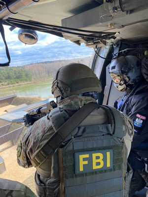 Pokład śmigłowca podczas lotu. Sfotografowany od tyłu funkcjonariusz FBI, policjant siedzi w otwartych drzwiach maszyny z wycelowanym w stronę tarczy strzeleckiej pistoletem maszynowym, niego obok Crew Chief.