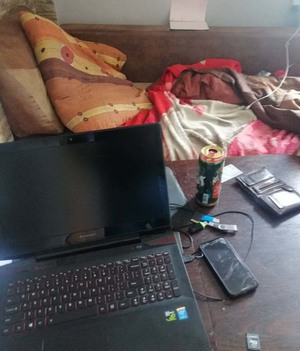 Stojący na stole laptop, leżący telefon, portfel i otwarta puszka piwa, z tyłu widoczne jest łóżko
