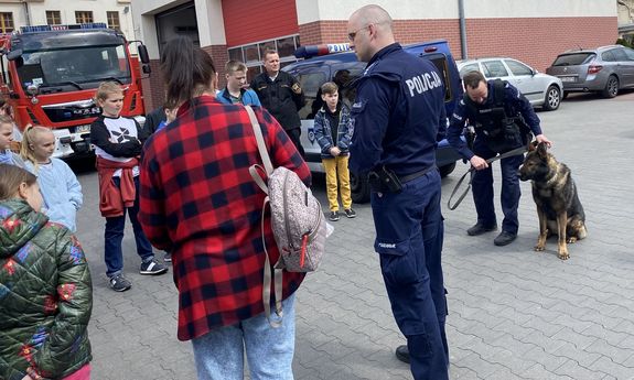 Policjanci, w tym jeden z policyjnym psem służbowym, stoją razem z grupą dzieci na terenie zewnętrznym jednostki pożarniczej