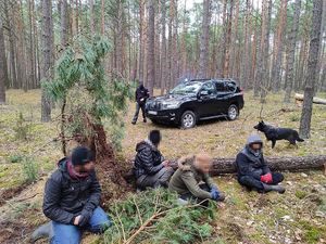 zdjęcie przedstawia czterech mężczyzn siedzących na ziemi w lesie w tle widać funkcjonariusza obok samochodu terenowego i psa