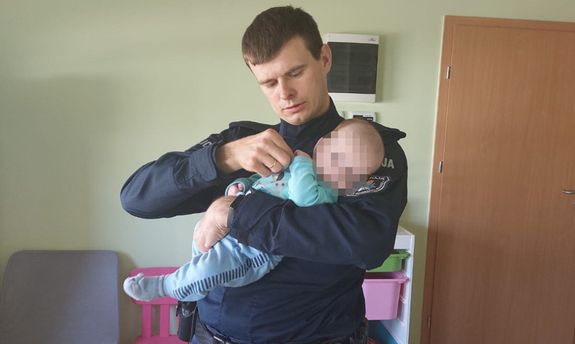 Zdjęcie przedstawia policjanta w granatowym mundurze, który trzyma w ramionach małe dziecko w niebieskich śpioszkach. Twarz dziecko jest zasłonięta komputerowym filtrem