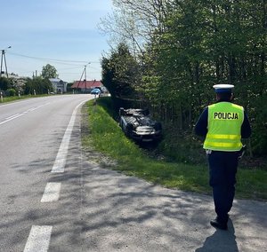 policjant ruchu drogowego stoi na poboczu drogi, w rowie widać przewrócony samochód leżący na dachu