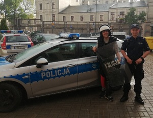 umundurowany policjant stoi z chłopcem w kasku z tarczą i kamizelką przy radiowozie