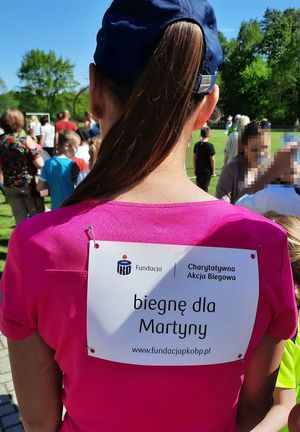 Dziewczyna w różowej bluzce stoi tyłem, na plecach ma kartkę z napisem Biegnę dla Martynki