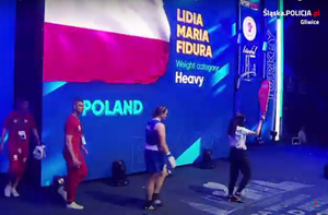 Trzy osoby w tym po środku Lidia fidura wchodzą na ring. W tle plansza z napisem POLAND Lidia Maria Fidura i flaga Polski