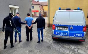 trzej policjanci z zatrzymanym mężczyzną, obok stoi radiowóz