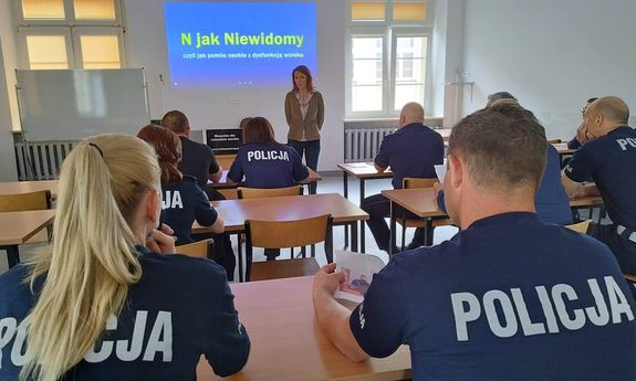 Zdjęcie wykonane zza pleców siedzących w ławkach szkolnych funkcjonariuszy Policji. W oddali prowadząca szkolenie a  na ścianie za nią napis „N jak Niewidomy, czyli jak pomóc osobie z dysfunkcja wzroku”