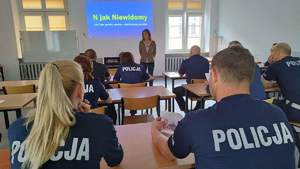 Zdjęcie wykonane zza pleców siedzących w ławkach szkolnych funkcjonariuszy Policji. W oddali prowadząca szkolenie a  na ścianie za nią napis „N jak Niewidomy, czyli jak pomóc osobie z dysfunkcja wzroku”