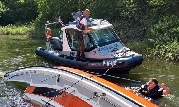 policjant na łodzi policyjnej, obok wywrócona żaglówka i drugi policjant w wodzie obok żaglówki