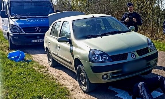 Policjant naprawia samochód. Leży pod samochodem. Z boku auta stoi drugi funkcjonariusz. Z tyłu widoczny jest policyjny furgon