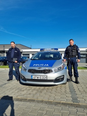 dwaj umundurowani policjanci stoją po obu stronach radiowozu