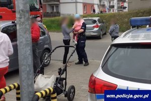 Miejsce interwencji. Na pierwszym planie wózek dziecięcy i radiowóz. W tle policjant z dzieckiem na ręku, a obok matka dziecka