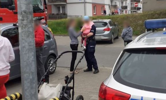 Miejsce interwencji. Na pierwszym planie wózek dziecięcy i radiowóz. W tle policjant z dzieckiem na ręku, a obok matka dziecka