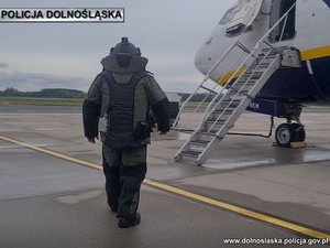 Policyjny pirotechnik w specjalistycznym kombinezonie idzie po płycie lotniska w stronę stojącego samolotu pasażerskiego