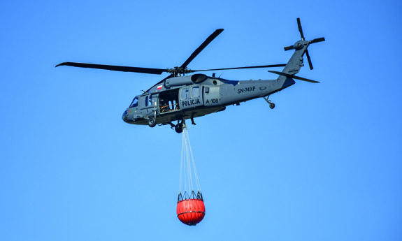 Policyjny Black Hawk z podwieszonym zbiornikiem na wodę do gaszenia pożarów.