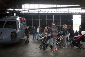 Przed stojącym w hangarze policyjnym Black Hawkiem. Grupa odwiedzających, pośród nich dzieci na wózkach spacerowych. W centrum kadru policjant w uniformie pilota.