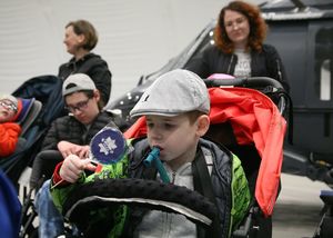 Trójka chłopców na wózkach. W centralnej części kadru jeden z nich wpatrzony w trzymany w ręku niebieski lizak, na którym znajduje się gwiazda Policji. W nieostrości tła dwie opiekunki i policyjny śmigłowiec Black Hawk.