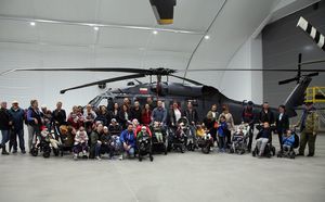 Zdjęcie pamiątkowe. We wnętrzu hangaru policyjnych śmigłowców Black Hawk sfotografowany z boku. Na jego tle grupa kilkunastu osób uczestniczących w wydarzeniu, pośród których dzieci na wózkach.