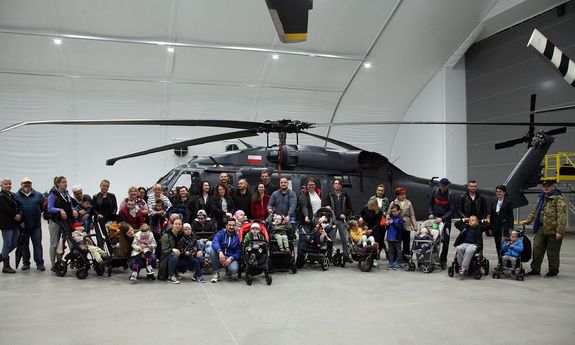 Zdjęcie pamiątkowe. We wnętrzu hangaru policyjnych śmigłowców Black Hawk sfotografowany z boku. Na jego tle grupa kilkunastu osób uczestniczących w wydarzeniu, pośród których dzieci na wózkach.