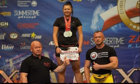 na najwyższym podium stoi kobieta, obok 2 meżczyźni