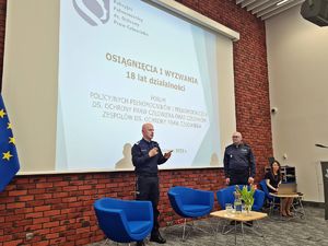 Dwaj umundurowani policjanci przemawiają na tle ekranu multimedialnego