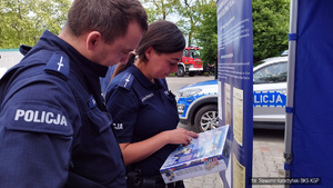Dwoje policjantów w mundurach ogląda grę planszowa pt. Zostań dzielnicowym na tle banneru Biuro Komunikacji Społecznej KGP. Na dalszych planach radiowóz policyjny i wóz bojowy Straży Pożarnej