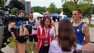 Pracownica BKS KGP w czerwonym dresie i koszulce reprezentacji Policji podczas wywiadu przed kamera telewizyjną. Za nią czterech policyjnych biegaczy w koszulkach z gwiazdą Policji na piersi