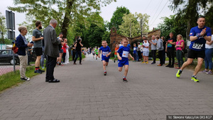 Kilkuletni zawodnicy w niebieskich strojach podczas biegu, za nimi kolejni, a na brzegach trasy klaszczący kibice