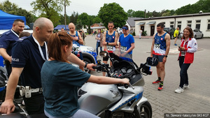 Niepełnosprawna uczestniczka biegu siedzi na policyjnym motocyklu, przy niej funkcjonariusz Policji w mundurze. W oddali policyjni lekkoatleci i kobieta w biało - czerwonym dresie