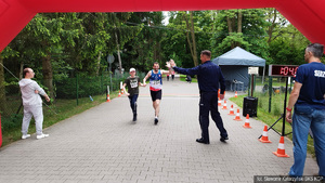 Linia mety. Policjant w mundurze „przybija piątkę” z biegaczem asystującym niepełnosprawnemu. Na elektronicznym zegarze czas do końca biegu 1.04.00