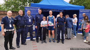 Policjanci przed stanowiskiem promocyjnym polskiej Policji. Pięcioro z nich jest w mundurach, jeden w stroju sportowym. W centrum kadru podinspektor trzyma rękę na ramieniu stojącego przed nim nastoletniego, niepełnosprawnego zawodnika.