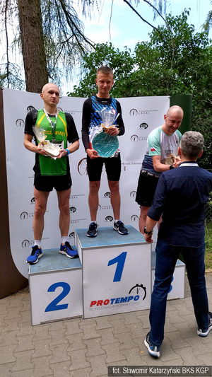 Podium a na nim na 1 miejscu policyjny lekkoatleta, na niższych stopniach podestu dwaj mężczyźni. Cała trójka z założonymi na szyjach medalami trzyma w rękach nagrody