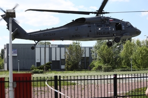 Black Hawk podczas podchodzenia do lądowania na utwardzonym placu. Z okna maszyny wychyla się „crew chief”. Na pierwszym planie - metalowy płot, w tle industrialny szary budynek.