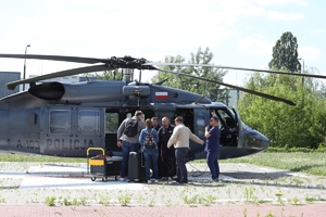 Słoneczny dzień, plac otaczają zielone drzewa, w pobliżu budynki. Na płycie lądowiska stoi policyjny Black Hawk z wyłączonym silnikiem. Przed otwartymi drzwiami ładowni troje osób po cywilnemu, lekarz i dwóch policyjnych lotników. Obok nich - na wózku transporter z organem do transplantacji.