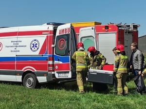 Trzej strażacy wkładają transporter z organem do wnętrza ambulansu Pogotowia Ratunkowego, funkcjonariuszom asystują dwie osoby w ubraniach cywilnych. Na dalszym planie wóz bojowy Straży Pożarnej.