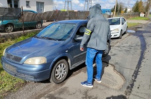 policjant z opaską na ręku z napisem Policja stoi przy ulicy obok niego zaparkowane dwa auta niebieskie i białe