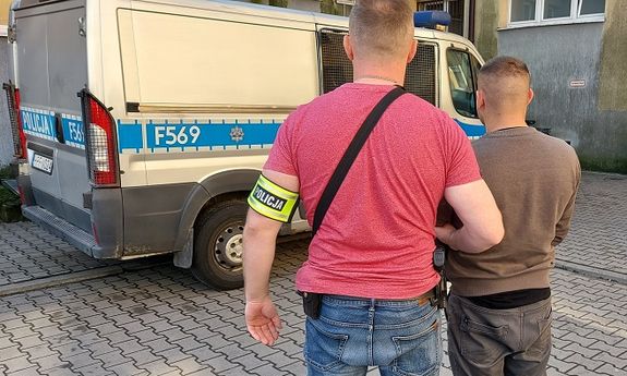 dziedziniec jednostki policjant z opaską na ręku z napisem Policja prowadzi zatrzymanego, w tle widać radiowóz