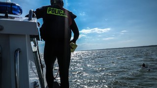 Mężczyzna w czarnym kombinezonie pływackim z pianki stoi na brzegu łodzi. Ma na sobie czarną kamizelkę z napisem Policja oraz kask.