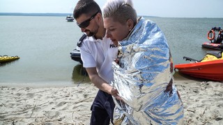 Funkcjonariusz Policji prowadzi kobietę zawinięta w srebrna folię zabezpieczającą przed utratą ciepła. Kilka metrów dalej stoją zaparkowane w wodzie łodzie.