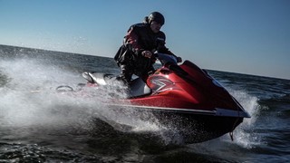 Mężczyzna w pozycji stojącej płynie z dużą prędkością na czerwonym skuterze wodnym. Ubrany jest w kurtkę z napisem Policja oraz kask.